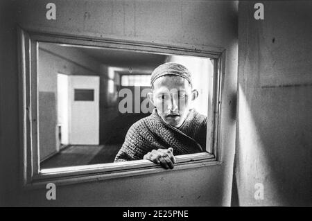Leninski Pyschiatric Hospital, Duschanbe, Tadschikistan. März 1998. Ein Mann, der an extremer PTBS leidet, schaut durch ein kaputtes Türfenster.1996 starben im größten, nationalen Krankenhaus namens Leninski in der Hauptstadt Duschanbe 176 Menschen. Aber psychische Erkrankungen sind normalerweise kein Killer. Diese Menschen starben an viel banaleren Ursachen: Hypothermie, Hunger, Lungenentzündung. Das staatliche Gesundheitssystem in Tadschikistan, das bis zur Unabhängigkeit 1991 von anderen Teilen der Sowjetunion stark subventioniert wurde, war zerfallen. Die Grundprioritäten in einem Land, das durch den Bürgerkrieg weiter bankrott gegangen ist, waren die psychiatrischen Einrichtungen Stockfoto