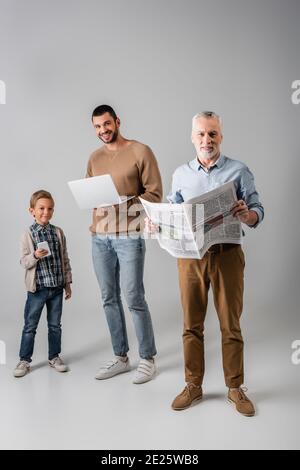 Reifer Mann hält Zeitung in der Nähe Sohn und Enkel mit Gadgets Auf Grau Stockfoto