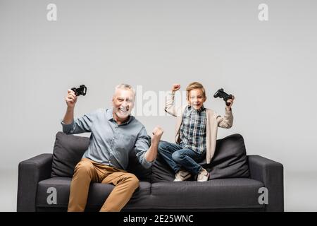 KIEW, UKRAINE - 17. NOVEMBER 2020: Aufgeregter Junge mit Opa zeigt Siegesthetus mit Joysticks isoliert auf grau Stockfoto