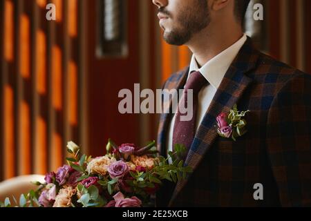 Das Porträt eines unerkennbaren, gutaussehenden Mannes in einer Krawatte und einem eleganten karierten Anzug mit Boutonniere, der einen großen Strauß frischer Blumen hält Stockfoto