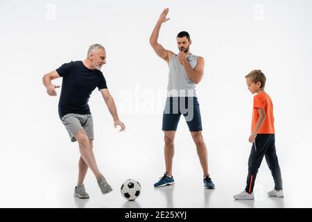 Mann in Sportkleidung pfeift in der Nähe von Großvater Fußball mit Enkel spielen Auf Weiß Stockfoto