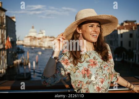 Lächelnde elegante Frau in Blumenkleid mit Hut zu erkunden Sehenswürdigkeiten auf der Accademia Brücke in Venedig, Italien. Stockfoto