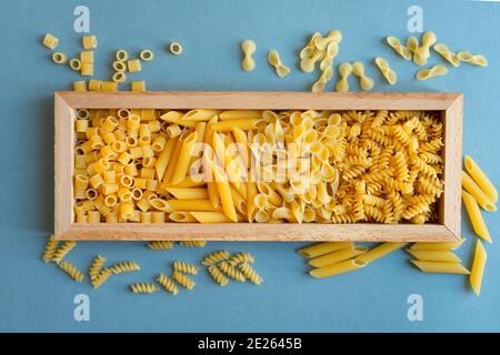 penne, farfalle tonde Schmetterling, Schnecke und Ditalini Art von Pasta auf hellblauem Hintergrund. Pasta in einer Holzkiste Teller. Abstrakt, Pasta. Draufsicht Pho Stockfoto