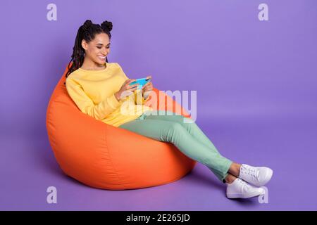 Foto in voller Größe von optimistischen Mädchen liegen auf Puff spielen Telefon tragen gelb Hemd Hose Sneakers isoliert auf lila Farbe Hintergrund Stockfoto