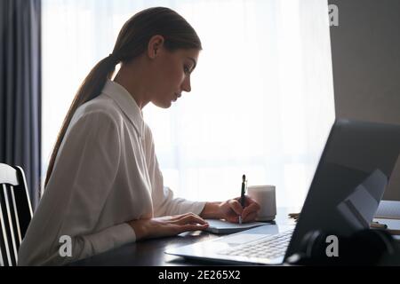 Seitenansicht der charmanten Dame, die ferngesteuerte Arbeit auf einem kabellosen Laptop macht, während sie am Tisch sitzt. Freiberuflerin in weißem Hemd mit tragbaren Computer bei ho Stockfoto