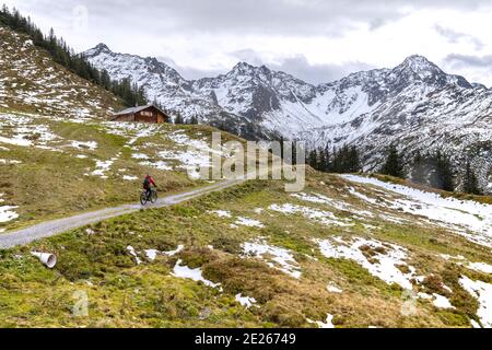 Zwei Mountainbiker, die im Herbst in Montafon, Vorarlberg, Österreich, auf dem Weg vorbei an Holzhütte auf Berghang mit schmelzenden Schneeflecken reiten Stockfoto