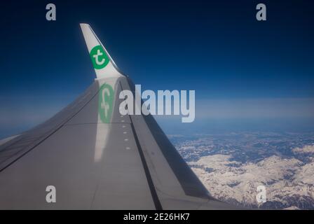 Transavia Flugzeug fliegt über schneebedeckten Bergen (Blick vom Fenster auf Flügel und Landschaft). Die Fluggesellschaft Transavia ist Teil der Air France-KLM-Gruppe. Stockfoto