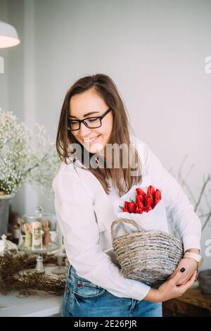 Schöne junge Mädchen in einem weißen Hemd hält einen Korb mit roten Tulpen unter dem Blumenladen. Frühlingskonzept, Blumensträuße aus frischen Blumen Stockfoto