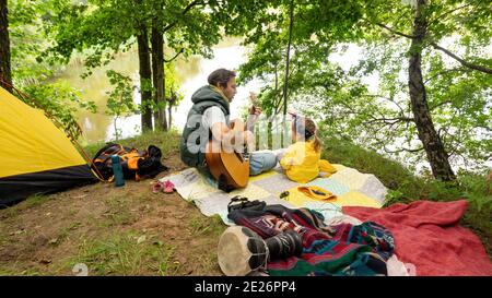 Ein Vater spielt für seine kleine Tochter Gitarre, während er auf dem Familiencamping am Ufer eines Sees Urlaub macht. Ruhe während der touristischen Trekking um Stockfoto