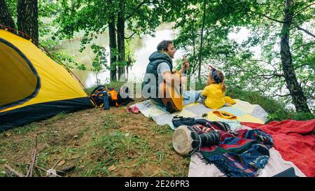 Vater und seine kleine Tochter sitzen im Wald auf dem Familiencamping und haben Spaß. Der Vater spielt für seine Tochter Gitarre. Ein Mann Tee Stockfoto