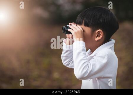 Kleiner asiatischer Junge, der durch ein Fernglas im Wald schaut. Das Kind beobachtete Ferngläser in der Natur im Freien. Stockfoto