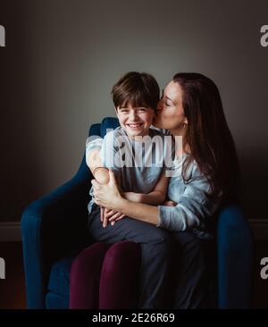 Frau umarmt und küsst ihren Sohn, während er in ihrem Schoß auf einem Stuhl sitzt. Stockfoto