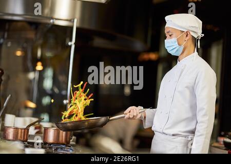 Talentierter asiatischer Koch schüttelt eine Pfanne mit Pfefferscheiben hinein Es beim Braten auf dem Herd Stockfoto