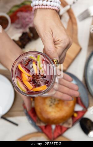 Die Hände einer Frau halten ein Glas Glühwein mit Zimt, geschnittener Frucht auf dem Hintergrund eines gedeckten Tisches. Der Blick von oben. Stockfoto