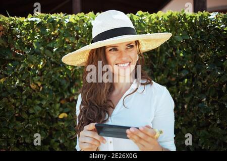 Lächelnd trendige Frau in weißem Hemd mit Hut mit Smartphone-Anwendungen im Freien in der Nähe von grünen Wand. Stockfoto