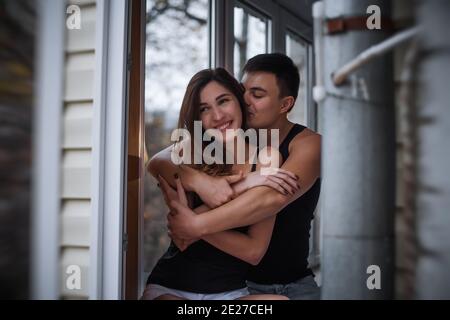 Ein liebevolles junges Paar umarmt sich im Fenster ihrer Balkonterrasse in der Wohnung. Der junge Mann lacht mit einer schönen Frau zu Hause. Liebhaber küssen, Pass