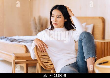 20s junge Teenager asiatische Frau trägt einen Pullover Blick weg in stilvollen gemütlichen Raum Hintergrund. Attraktive weibliche Modell posiert für Kamera auf Stuhl. Winter Portrait Mode Lifestyle. Platz für die Banner-Kopie Stockfoto