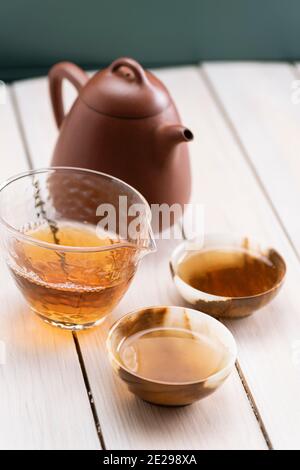 Gesundes traditionelles Getränkekonzept. Chinesischer alter shen puerh Tee in Ton Teekanne gebrüht und serviert in Glas Chahai und Onyx Steinbecher auf weißem Holz Stockfoto