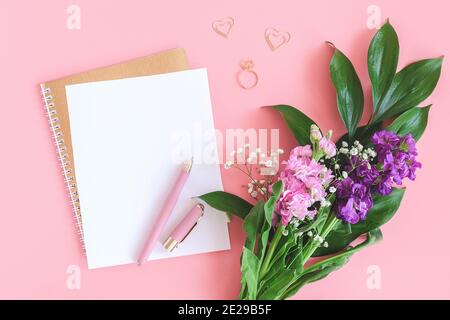 Weißes Blatt auf spiralförmigem goldenem Notizblock mit Büroklammer Herz und Ring und Blumenstrauß auf rosa Hintergrund. Konzept romantische Liebe Mockup für Stockfoto