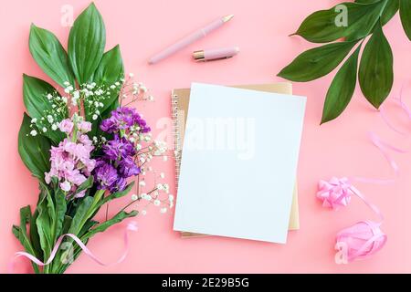 Blankes weißes Blatt auf spiralförmigem goldenem Notizblock mit Stift für Ihren Text oder Ihr Design und Blumenstrauß auf rosa Hintergrund. Konzept weiblicher Arbeitsbereich Mockup Stockfoto