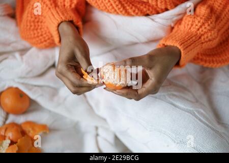 Afro Frau Hände schälen reife süße Mandarine, tragen orange Pullover, liegen im Bett unter dem Plaid Stockfoto