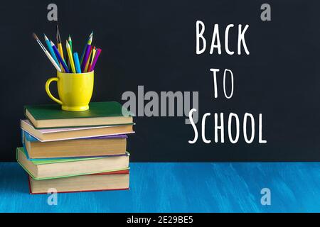Zurück zur Schule Text auf schwarzer Tafel und Schreibwaren in einem gelben Becher auf Stapel von Büchern auf blauem Tisch. Konzeptbildung. Vorderansicht. Stockfoto
