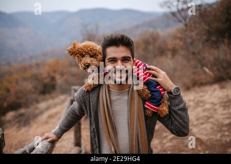 Mann steht und hält Hund auf Schultern in der Natur. Herbstzeit. Im Hintergrund Berge und Wald. Stockfoto