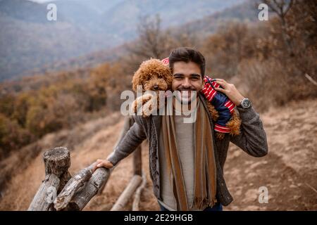 Mann steht und hält Hund auf Schultern in der Natur. Herbstzeit. Im Hintergrund Berge und Wald. Stockfoto