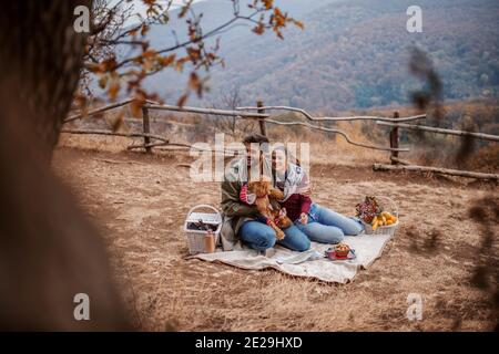 Pärchen sitzt auf der Decke beim Picknick mit ihrem Hund. Daneben Obstkorb und Picknickkorb. Herbstzeit, im Hintergrund Berg. Stockfoto