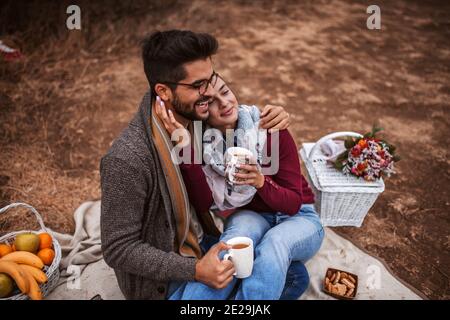 Frau, die beim Picknick im Schoß des Mannes sitzt. Paar kuscheln und Tee trinken. Daneben Picknickkorb, Blumen und Obstkorb. Herbstzeit. Stockfoto