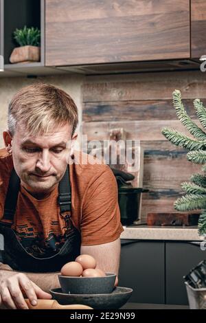 Kaukasischer Bäcker in einer Schürze in einer Wohnküche. Mann kochen zu Hause. Lifestyle-Fotografie. Vertikale Aufnahme. Hausarbeit. Vertikale Aufnahme Stockfoto