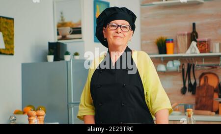 Alte Dame trägt Chef Schürze und bonete in der Küche mit Mehl auf dem Tisch zum Backen Blick auf Kamera und Lächeln. Rentner älterer Bäcker, der Backzutaten zubereitet, um hausgemachtes Brot und Kuchen zu kochen Stockfoto