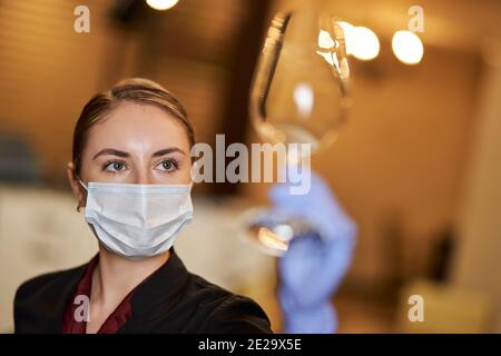 Aufmerksame junge Frau in Schutzmaske Blick eng auf ein Klares Weinglas in der Hand Stockfoto