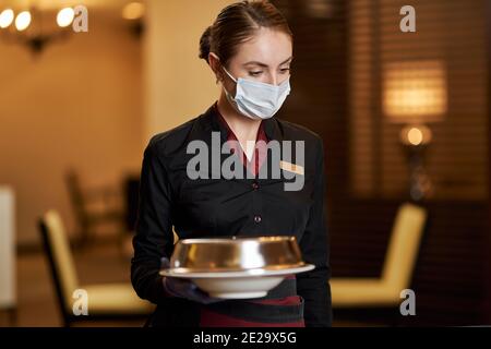 Serious junge Frau in Corporate Uniform und Gesichtsmaske tragen Eine Platte mit Deckel bedeckt Stockfoto