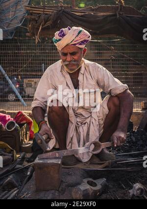 Jaisalmer, Rajasthan / Indien - 9. Januar 2021 : alter indischer Schmied trägt Turban Schmieden ein Stück Metall in seiner kleinen Straße Straße Straße Werkstatt Stockfoto