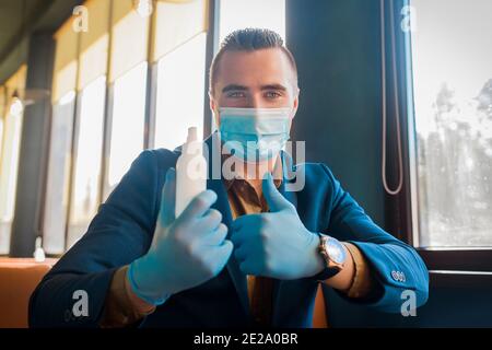 Junge kaukasische männliche Geschäftsmann in einer Schutzmaske und medizinischen Handschuhen hält ein Antiseptikum und zeigt Daumen nach oben. Stockfoto