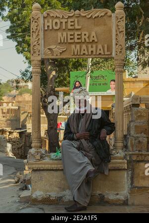 Jaisalmer, Rajasthan / Indien - 8. Januar 2021 : Alter stilvoller Mann trägt Turban und unsachgemäße Gesichtsmaske sitzt unter einer Plakatwand aus Stein geschnitzt Stockfoto