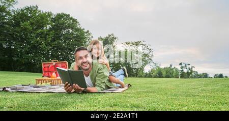 Fröhliche niedliche kleine Mädchen verbringen Zeit mit ihrem Vater, ein Buch zu lesen, während mit Picknick im Park Stockfoto