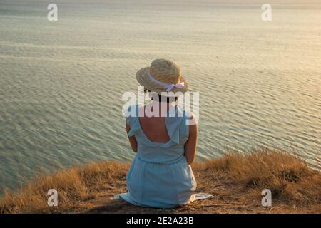 Ein Mädchen in einem Kleid und Hut sitzt auf einem Klippe am Meer Stockfoto