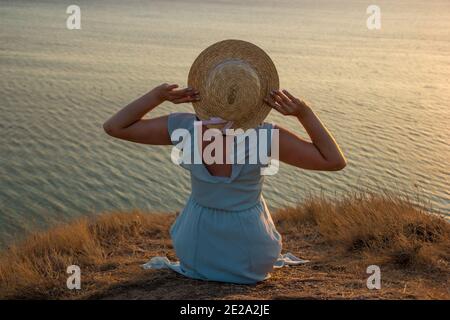 Ein Mädchen in einem Kleid und Hut sitzt auf einem Klippe am Meer Stockfoto