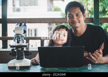 Lächelnder Vater und Tochter lernen von zu Hause aus mit Laptop und Microscope.Coronavirus oder Covid-19 Ausbruch Schule Abschaltungen Stockfoto
