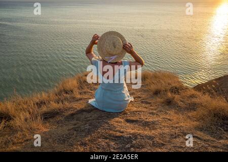 Ein Mädchen in einem Kleid und Hut sitzt auf einem Klippe am Meer Stockfoto