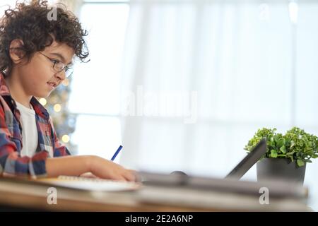 Begeisterter lateinamerikanischer Schuljunge, der mit einer Brille Hausaufgaben mit einem digitalen Tablet macht und zu Hause am Schreibtisch sitzt. Online-Bildung, Technologie, Homeschooling. Seitenansicht Stockfoto