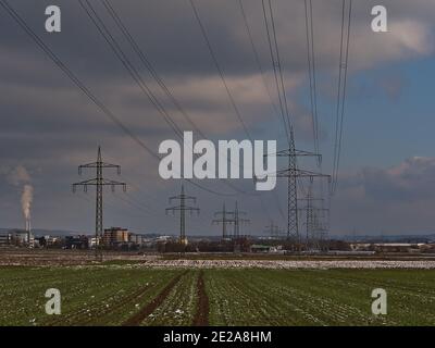 Elektrische Stromleitungen mit großen Metallmasten über grünen landwirtschaftlichen Feldern in der Wintersaison mit Restschnee an bewölkten Tagen. Stockfoto