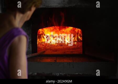 Feuer und Flammen sind in einem alten ländlichen Holzofen oder Ofen zu sehen. Eine Person, eine Frau, ist vor dem Ofen zu sehen. Stockfoto