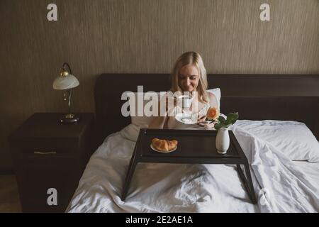Frühstück im Bett. Eine Frau im Hotel oder Zimmer trinkt Kaffee im Bett sitzend, vor ihr ist Tisch-Tablett mit Croissant und Vase mit Rose Stockfoto