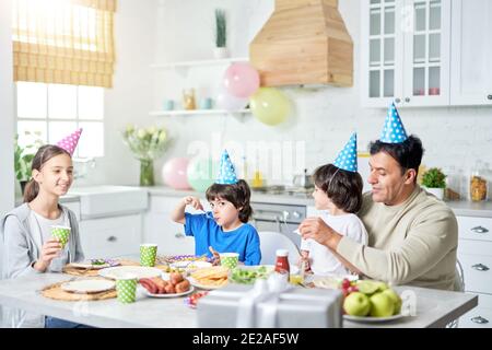 Liebevoller lateinischer Vater mittleren Alters mit Kindern, die Geburtstagsmützen auf den Köpfen tragen, und mit einem Abendessen, während wir zu Hause gemeinsam Geburtstag feiern. Vaterschaft, glückliches Familienkonzept Stockfoto