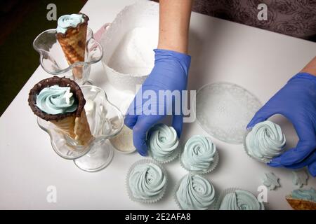 Frau in Gummihandschuhen macht Marshmallows. In der Nähe befindet sich ein zephyr in einem Waffelkegel. Dessert liegt auf der Tischoberfläche. Stockfoto