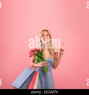 Glückliche junge Dame trägt schönes Kleid, hält Frühlingsblumen und Geschenktüten, zeigt auf leeren Raum auf rosa Hintergrund Stockfoto