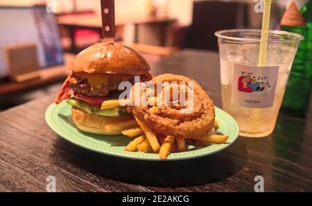 Ein Gourmet-Hamburger mit gebratenen Zwiebelringen, Pommes frites und einem Drink auf einem dunklen Holztisch in einem gut beleuchteten Restaurant Stockfoto